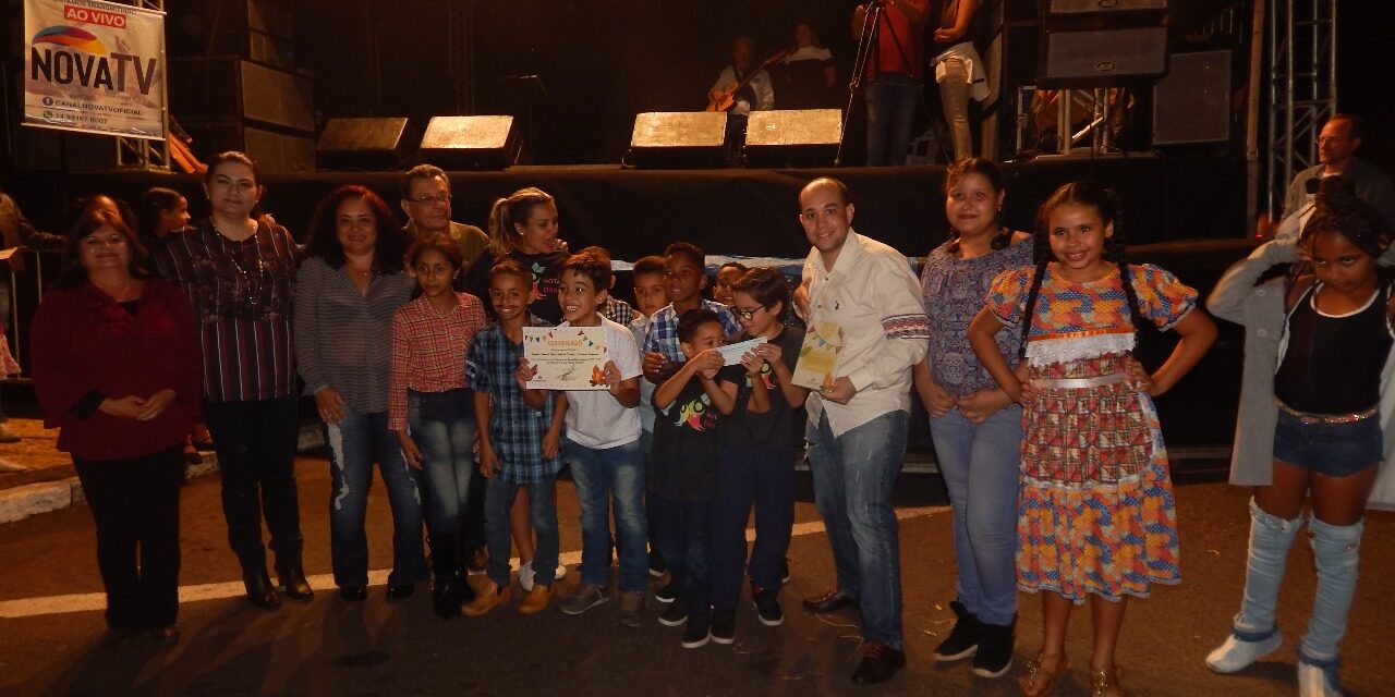 Solenidade noturna: Celebrando jovens talentos e parcerias. Grupo de crianças e adultos posa para foto noturna em palco iluminado, exibindo certificados e com a logo.
