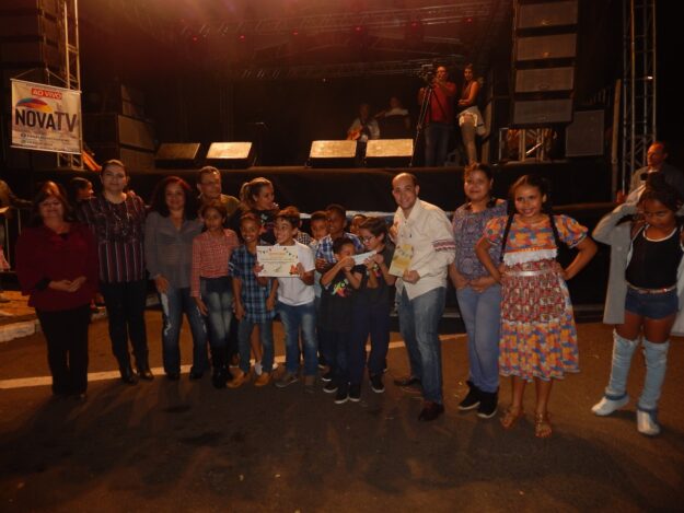 Solenidade noturna: Celebrando jovens talentos e parcerias. Grupo de crianças e adultos posa para foto noturna em palco iluminado, exibindo certificados e com a logo.