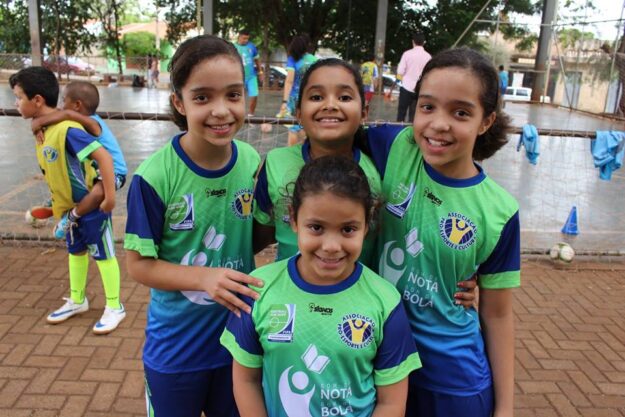 Futebol feminino: Amizade e união em campo. Quatro crianças jogadoras de futebol em uniformes verde e azul, sorrindo e abraçadas em quadra aberta de terra batida.