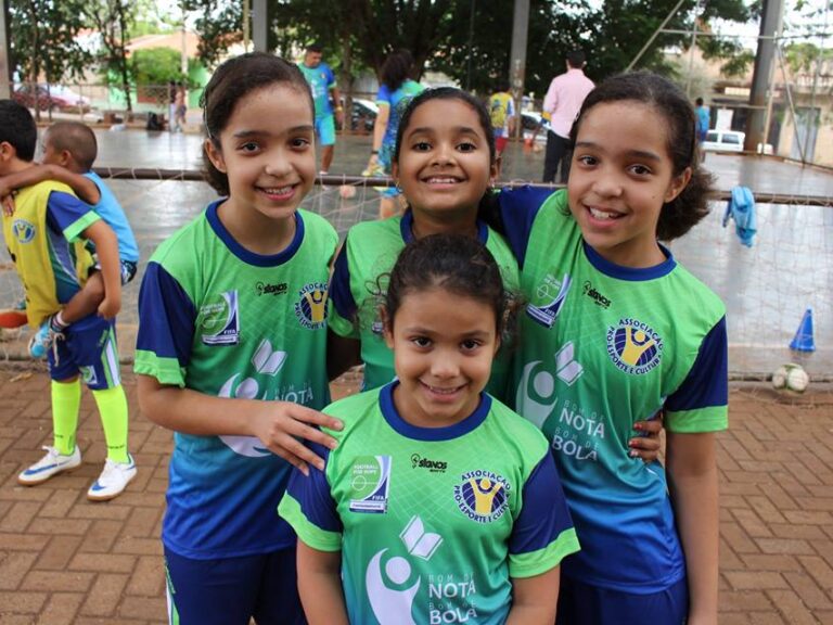 Futebol feminino: Amizade e união em campo. Quatro crianças jogadoras de futebol em uniformes verde e azul, sorrindo e abraçadas em quadra aberta de terra batida.