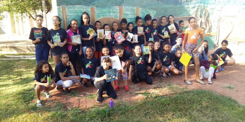 Incentivo à leitura em grupo: Jovens leitores ao ar livre. Grande grupo de crianças posa em área externa com um mural pintado, exibindo diversos livros e gibis em suas mãos.