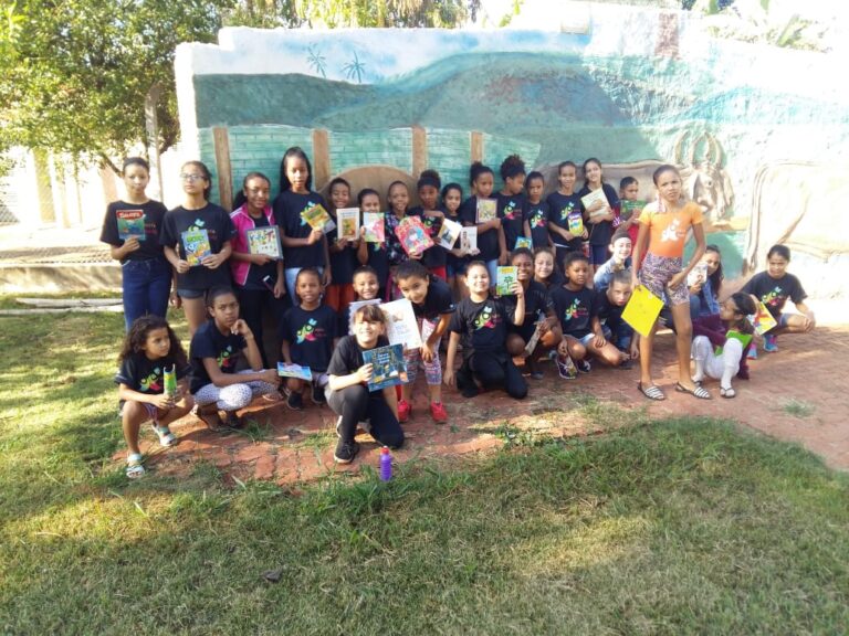 Incentivo à leitura em grupo: Jovens leitores ao ar livre. Grande grupo de crianças posa em área externa com um mural pintado, exibindo diversos livros e gibis em suas mãos.