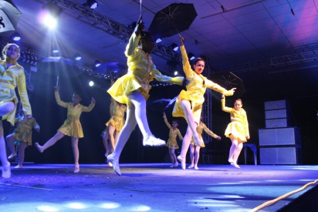 Espetáculo de Dança. Dançarinas em trajes de chuva amarelos e guarda-chuvas pretos se apresentam no palco com luzes de LED azul e branca.
