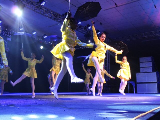 Espetáculo de Dança. Dançarinas em trajes de chuva amarelos e guarda-chuvas pretos se apresentam no palco com luzes de LED azul e branca.