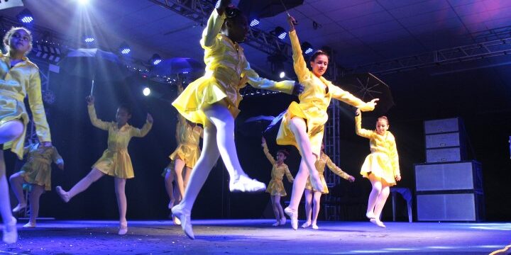 Espetáculo de Dança. Dançarinas em trajes de chuva amarelos e guarda-chuvas pretos se apresentam no palco com luzes de LED azul e branca.