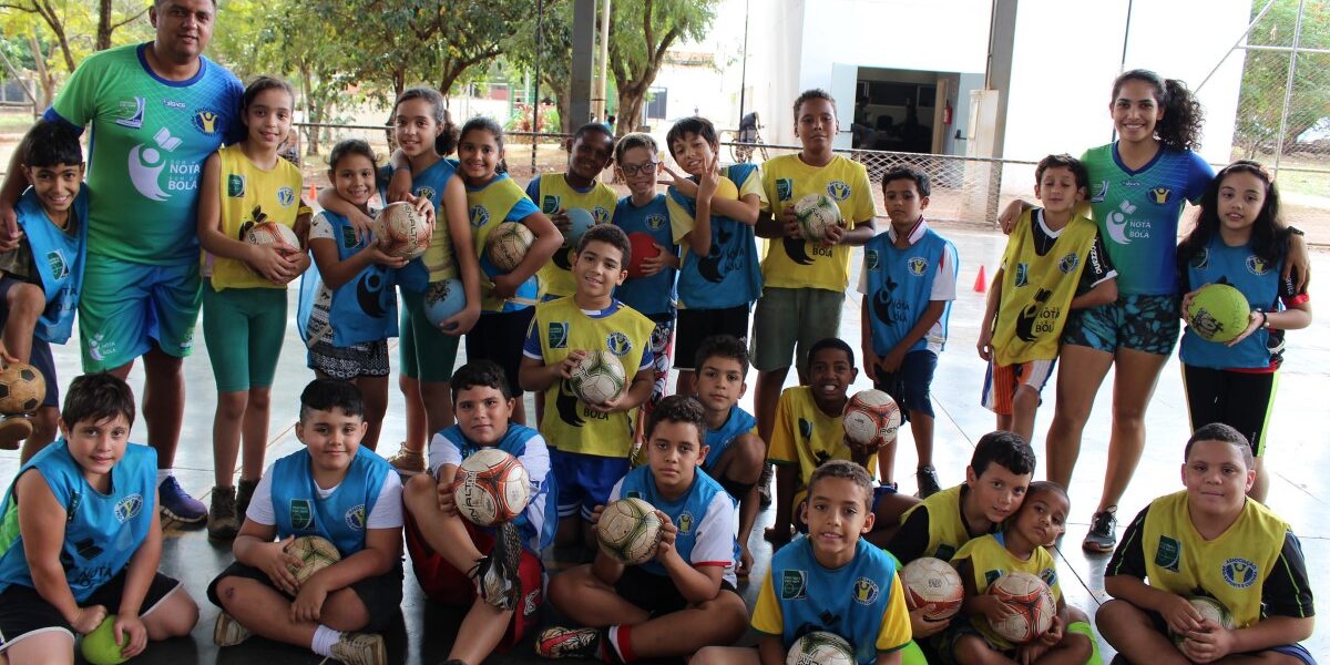 Time de futebol no treinamento. Grande grupo de crianças e dois adultos em quadra coberta. Todos vestem uniformes esportivos e seguram bolas de futebol.