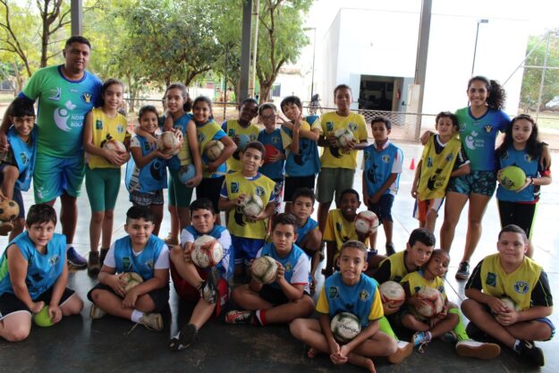 Time de futebol no treinamento. Grande grupo de crianças e dois adultos em quadra coberta. Todos vestem uniformes esportivos e seguram bolas de futebol.