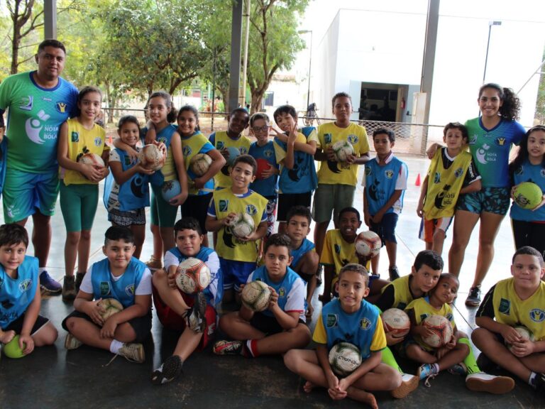Time de futebol no treinamento. Grande grupo de crianças e dois adultos em quadra coberta. Todos vestem uniformes esportivos e seguram bolas de futebol.