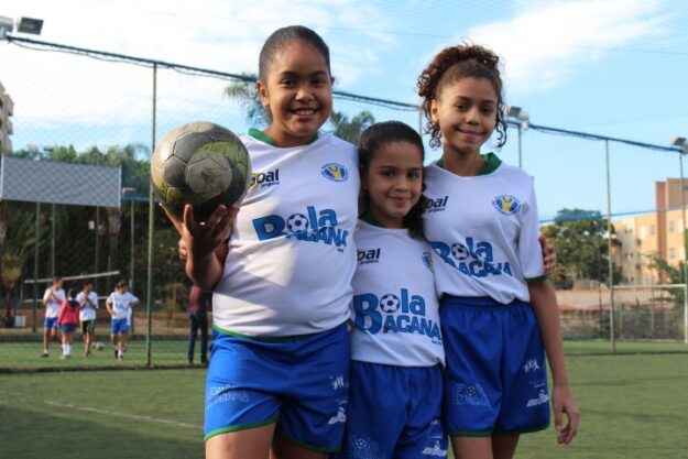 Futebol feminino juvenil: alegria no campo. Três crianças jogadoras de futebol em uniformes brancos e azuis, sorrindo em campo de grama sintética. Uma delas segura uma bola.
