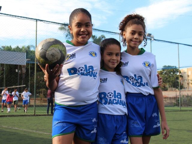 Futebol feminino juvenil: alegria no campo. Três crianças jogadoras de futebol em uniformes brancos e azuis, sorrindo em campo de grama sintética. Uma delas segura uma bola.