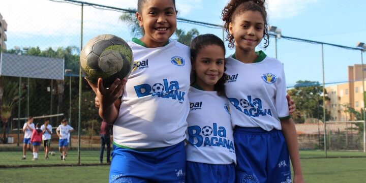 Futebol feminino juvenil: alegria no campo. Três crianças jogadoras de futebol em uniformes brancos e azuis, sorrindo em campo de grama sintética. Uma delas segura uma bola.