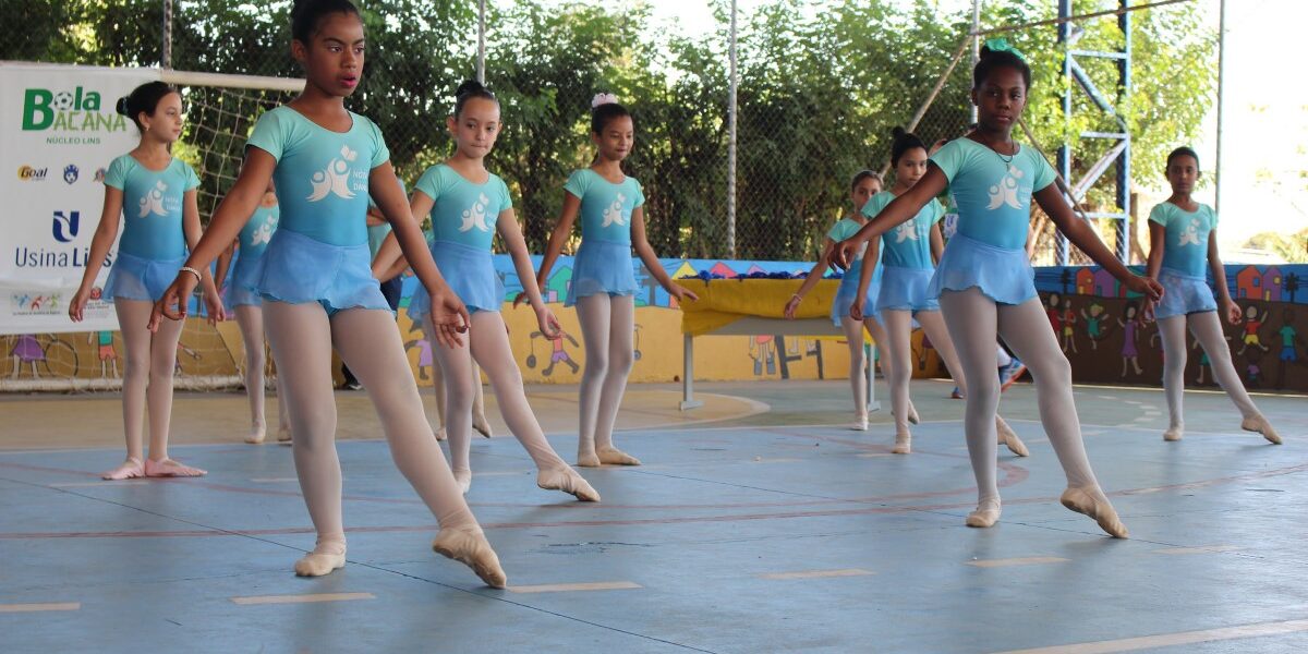 Apresentação de balé em quadra poliesportiva. Jovens alunas de balé se apresentam em quadra esportiva, vestindo collants azuis claros e meia-calça. O fundo tem uma tela de patrocinadores.