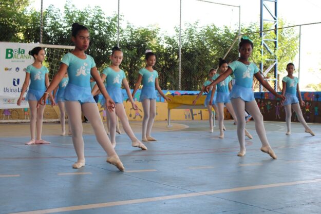 Apresentação de balé em quadra poliesportiva. Jovens alunas de balé se apresentam em quadra esportiva, vestindo collants azuis claros e meia-calça. O fundo tem uma tela de patrocinadores.