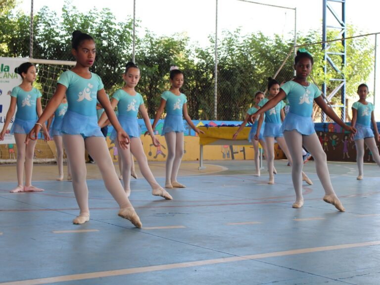 Apresentação de balé em quadra poliesportiva. Jovens alunas de balé se apresentam em quadra esportiva, vestindo collants azuis claros e meia-calça. O fundo tem uma tela de patrocinadores.