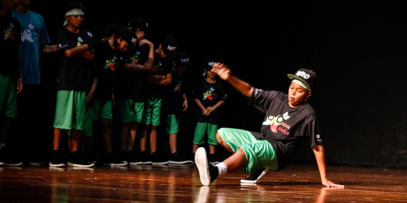 Performance de breakdance no palco. Jovem dançarino de breakdance em camiseta preta e bermuda verde, realizando um movimento no palco escuro. Outras crianças observam.