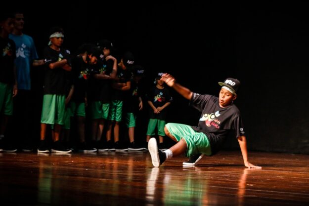 Performance de breakdance no palco. Jovem dançarino de breakdance em camiseta preta e bermuda verde, realizando um movimento no palco escuro. Outras crianças observam.