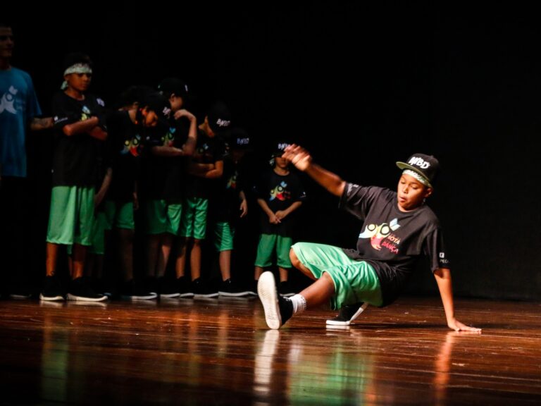 Performance de breakdance no palco. Jovem dançarino de breakdance em camiseta preta e bermuda verde, realizando um movimento no palco escuro. Outras crianças observam.