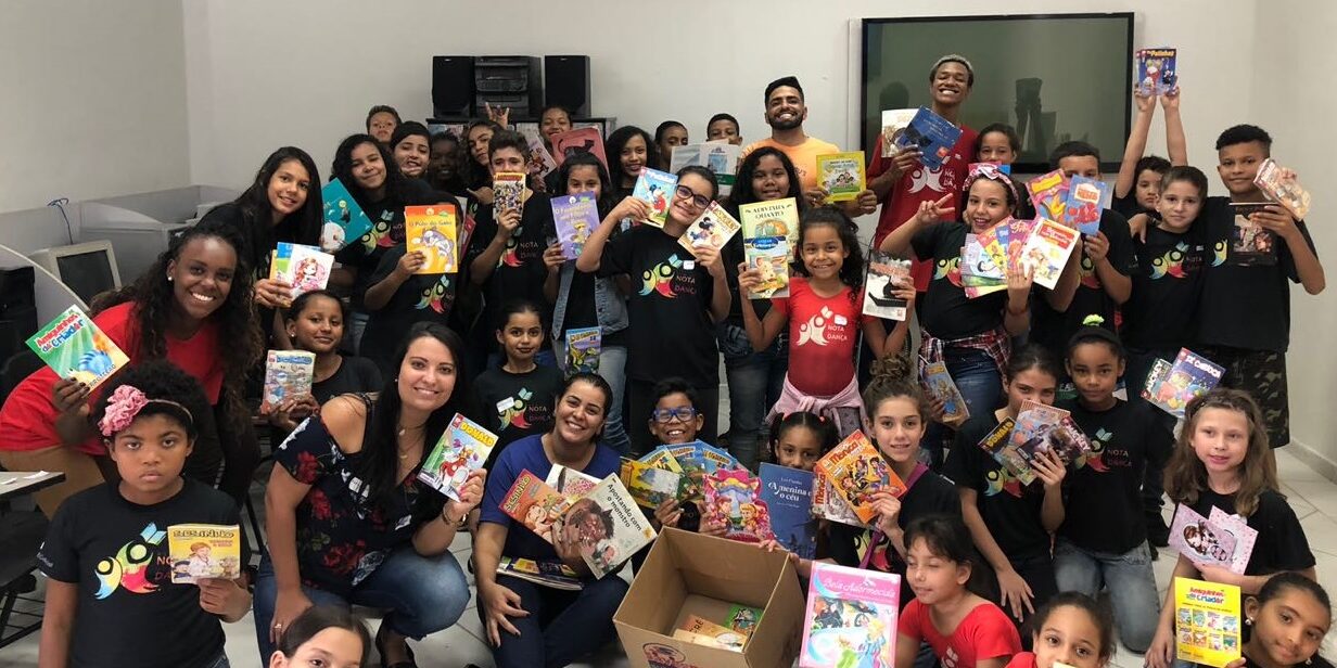 Celebração de doação de livros e leitura infantil. Grande grupo de crianças e adultos em sala, celebrando a doação de livros e gibis, segurando as publicações alegremente.