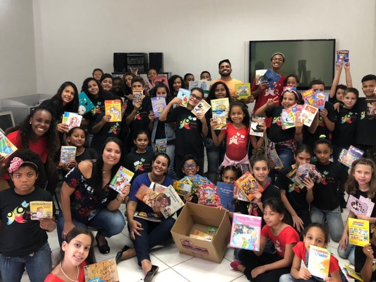 Celebração de doação de livros e leitura infantil. Grande grupo de crianças e adultos em sala, celebrando a doação de livros e gibis, segurando as publicações alegremente.