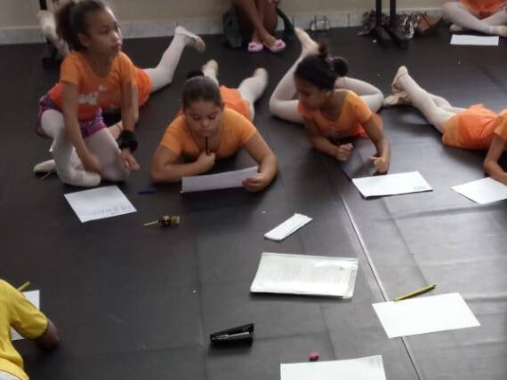 Momento de criação e foco no estúdio de dança. Crianças vestindo collants de dança e roupas esportivas, deitadas e sentadas no chão, concentradas escrevendo em papéis.