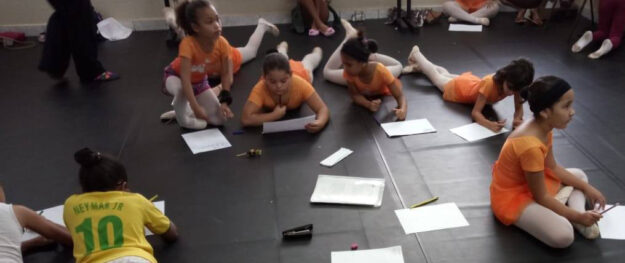 Momento de criação e foco no estúdio de dança. Crianças vestindo collants de dança e roupas esportivas, deitadas e sentadas no chão, concentradas escrevendo em papéis.