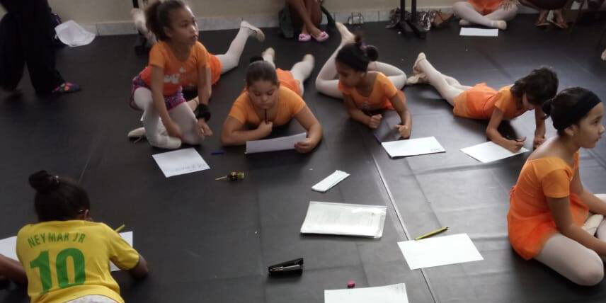 Momento de criação e foco no estúdio de dança. Crianças vestindo collants de dança e roupas esportivas, deitadas e sentadas no chão, concentradas escrevendo em papéis.