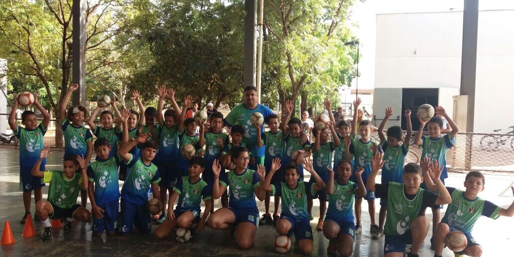 Time de futebol infantil: União e entusiasmo. Grande grupo de crianças e um adulto em quadra coberta. Todos vestem uniformes de futebol verde e azul e levantam os braços com bolas.