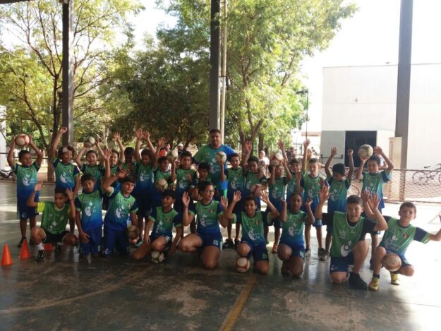 Time de futebol infantil: União e entusiasmo. Grande grupo de crianças e um adulto em quadra coberta. Todos vestem uniformes de futebol verde e azul e levantam os braços com bolas.