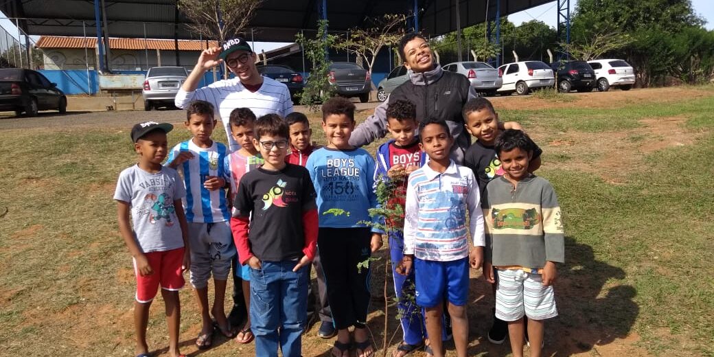 Plantio de mudas e conscientização ambiental com crianças. Grupo de dez crianças com dois adultos em área verde, celebrando o plantio de uma pequena muda de árvore no centro da foto.