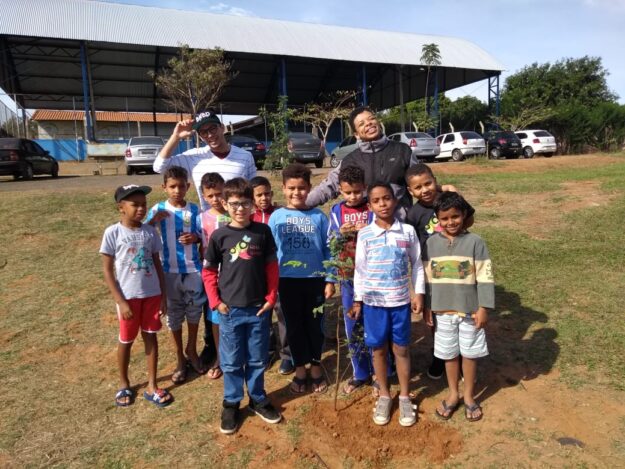 Plantio de mudas e conscientização ambiental com crianças. Grupo de dez crianças com dois adultos em área verde, celebrando o plantio de uma pequena muda de árvore no centro da foto.