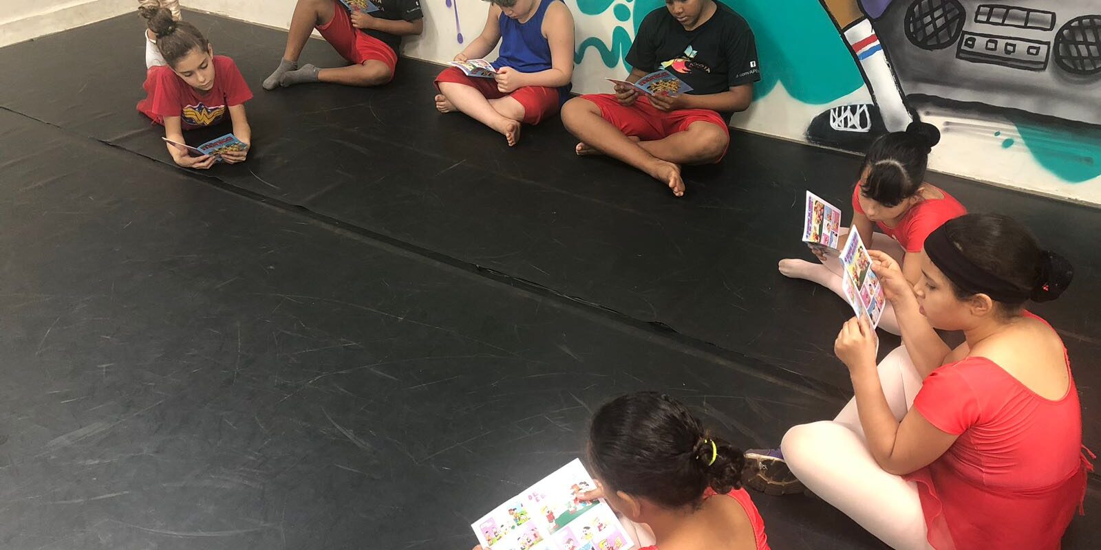 Momento de leitura em sala de arte urbana. Grupo de crianças sentadas no chão de um estúdio de dança, lendo gibis ou livros de imagens, com grafite ao fundo.