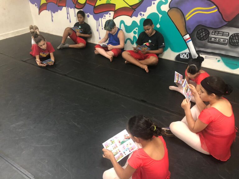 Momento de leitura em sala de arte urbana. Grupo de crianças sentadas no chão de um estúdio de dança, lendo gibis ou livros de imagens, com grafite ao fundo.