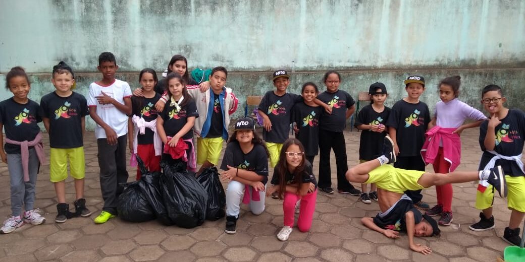 O Futuro em Ação: Crianças promovem a limpeza urbana. Grupo de crianças e jovens posa em pátio, com um deles realizando um movimento acrobático e sacos de lixo preto ao lado.