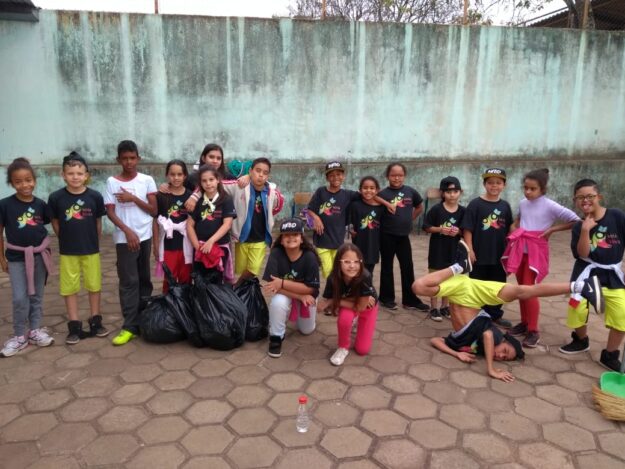 O Futuro em Ação: Crianças promovem a limpeza urbana. Grupo de crianças e jovens posa em pátio, com um deles realizando um movimento acrobático e sacos de lixo preto ao lado.