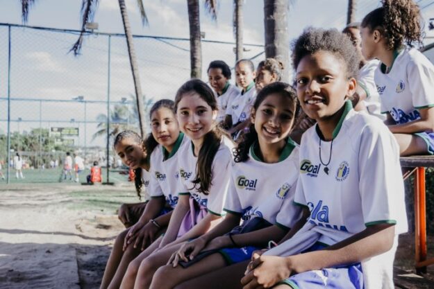 Empoderamento e união no esporte feminino. Jovens atletas em uniforme de projeto esportivo posam sorridentes. Outras crianças jogam futebol ao fundo.