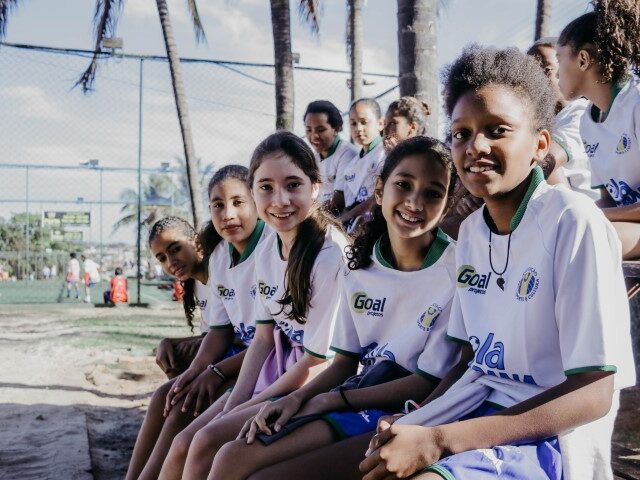 Empoderamento e união no esporte feminino. Jovens atletas em uniforme de projeto esportivo posam sorridentes. Outras crianças jogam futebol ao fundo.