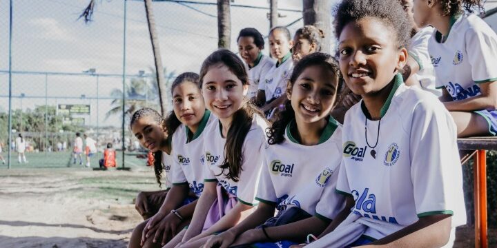 Empoderamento e união no esporte feminino. Jovens atletas em uniforme de projeto esportivo posam sorridentes. Outras crianças jogam futebol ao fundo.