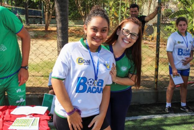 Alegria e participação no esporte social. Aluna e professora posam juntas em evento esportivo social, com camisa do projeto "Bola Bacana"
