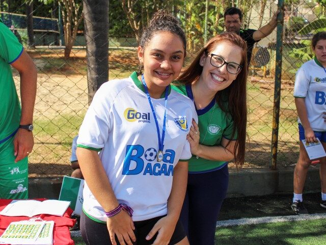 Alegria e participação no esporte social. Aluna e professora posam juntas em evento esportivo social, com camisa do projeto "Bola Bacana"