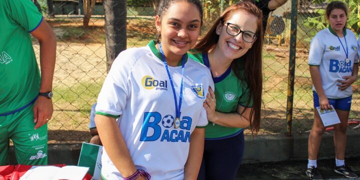 Alegria e participação no esporte social. Aluna e professora posam juntas em evento esportivo social, com camisa do projeto "Bola Bacana"