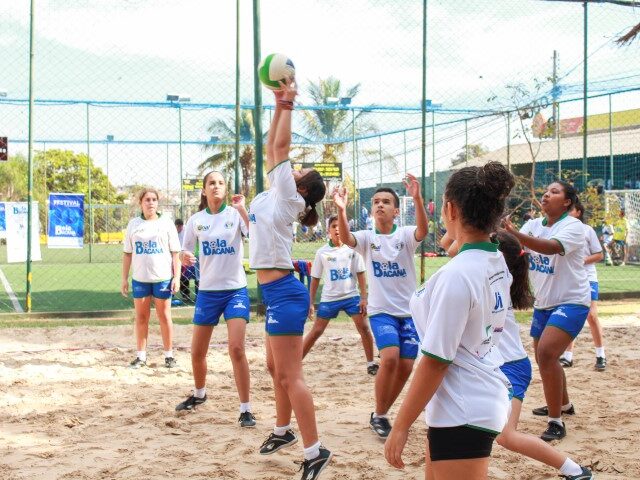Vôlei de areia juvenil em ação. Grupo de jovens em uniformes brancos e azuis jogando vôlei de areia, com uma atleta no centro preparando-se para sacar a bola.