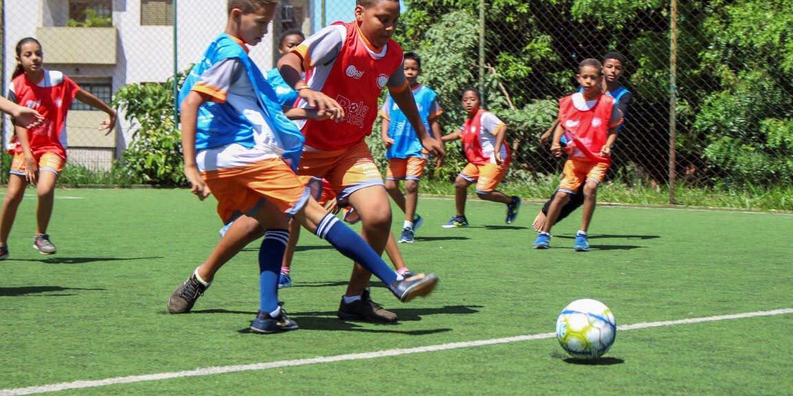 Partida infantil do projeto Bola Bacana Crianças jogando futebol em campo gramado, usando coletes vermelhos e azuis, concentradas na disputa pela bola.