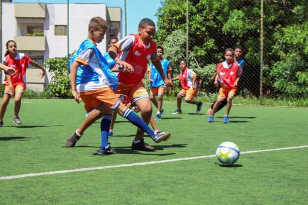 Partida infantil do projeto Bola Bacana Crianças jogando futebol em campo gramado, usando coletes vermelhos e azuis, concentradas na disputa pela bola.