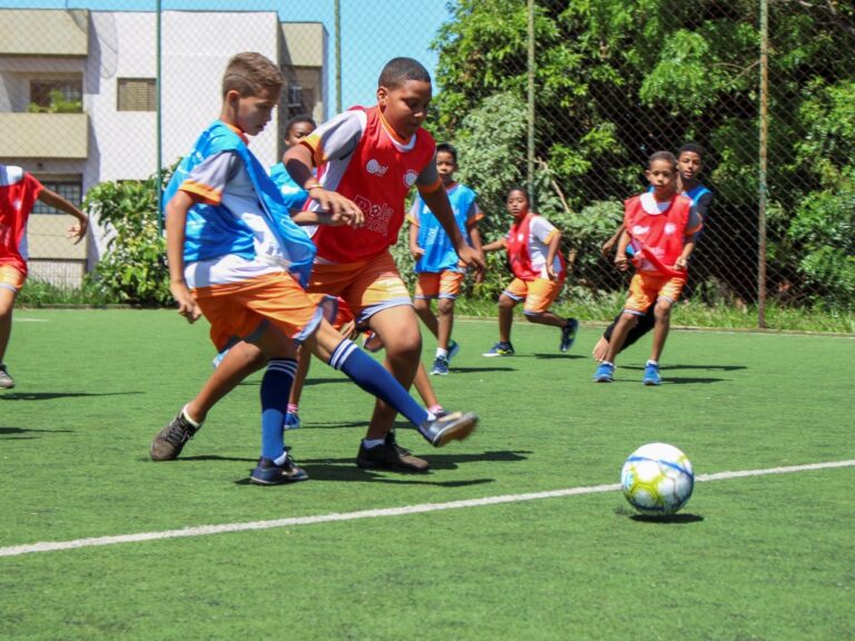 Partida infantil do projeto Bola Bacana Crianças jogando futebol em campo gramado, usando coletes vermelhos e azuis, concentradas na disputa pela bola.