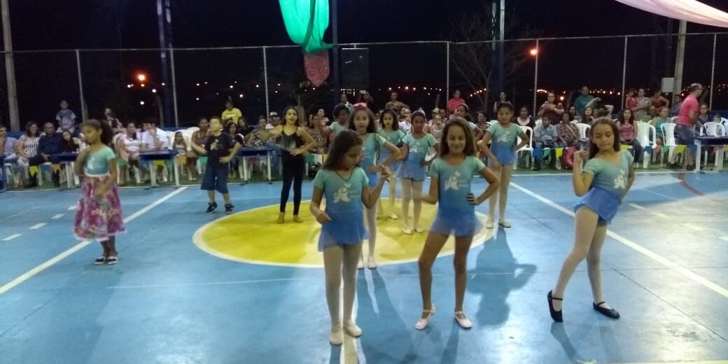 Espetáculo de dança infantil em noite festiva. Grupo de meninas em trajes de dança azul e branco, se apresentando em quadra esportiva com decoração de tecidos coloridos.