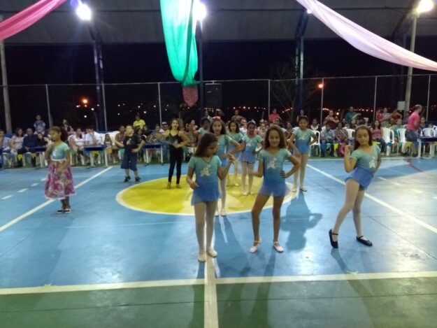 Espetáculo de dança infantil em noite festiva. Grupo de meninas em trajes de dança azul e branco, se apresentando em quadra esportiva com decoração de tecidos coloridos.