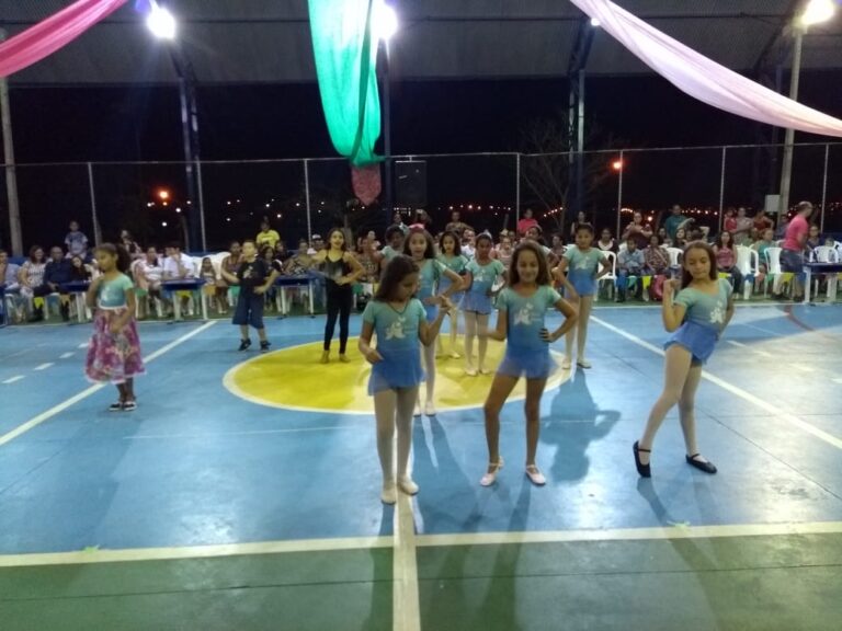 Espetáculo de dança infantil em noite festiva. Grupo de meninas em trajes de dança azul e branco, se apresentando em quadra esportiva com decoração de tecidos coloridos.