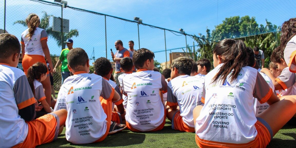 O esporte conectando crianças e oportunidades. Crianças sentadas em gramado sintético, de costas, vestindo camisetas com logos de patrocinadores e do Ministério do Esporte do Governo Federal, em um evento esportivo