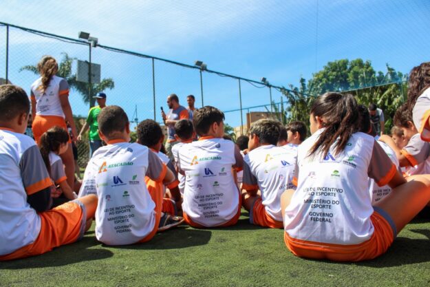 O esporte conectando crianças e oportunidades. Crianças sentadas em gramado sintético, de costas, vestindo camisetas com logos de patrocinadores e do Ministério do Esporte do Governo Federal, em um evento esportivo