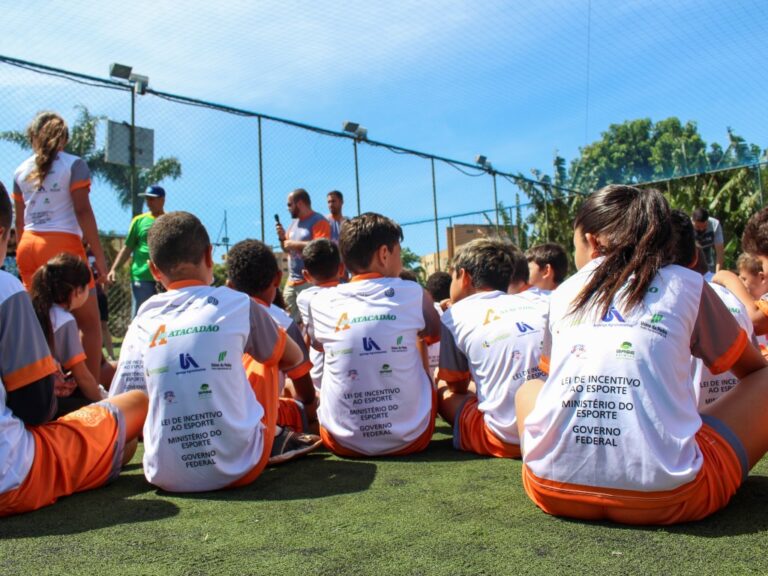 O esporte conectando crianças e oportunidades. Crianças sentadas em gramado sintético, de costas, vestindo camisetas com logos de patrocinadores e do Ministério do Esporte do Governo Federal, em um evento esportivo